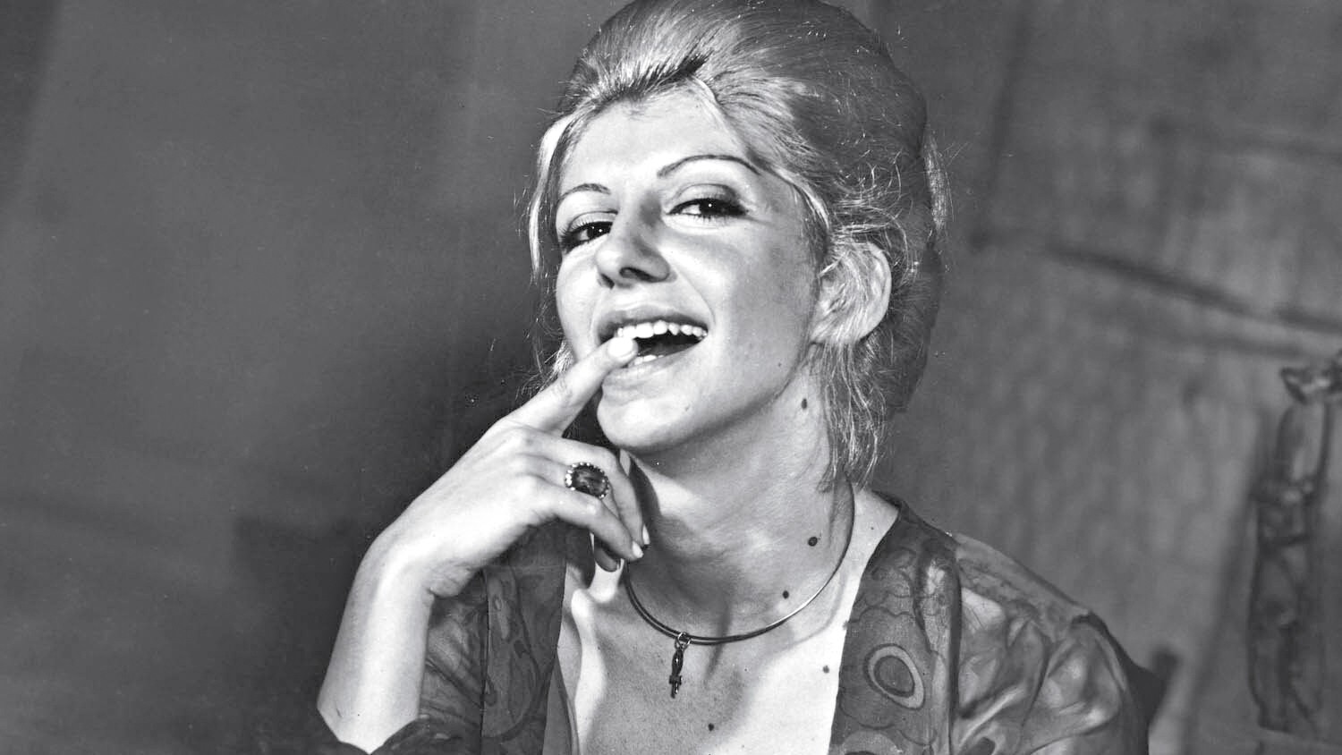 Mulher loira com cabelo volumoso, sorrindo e mordendo o dedo indicador, usando anel e colar, em preto e branco