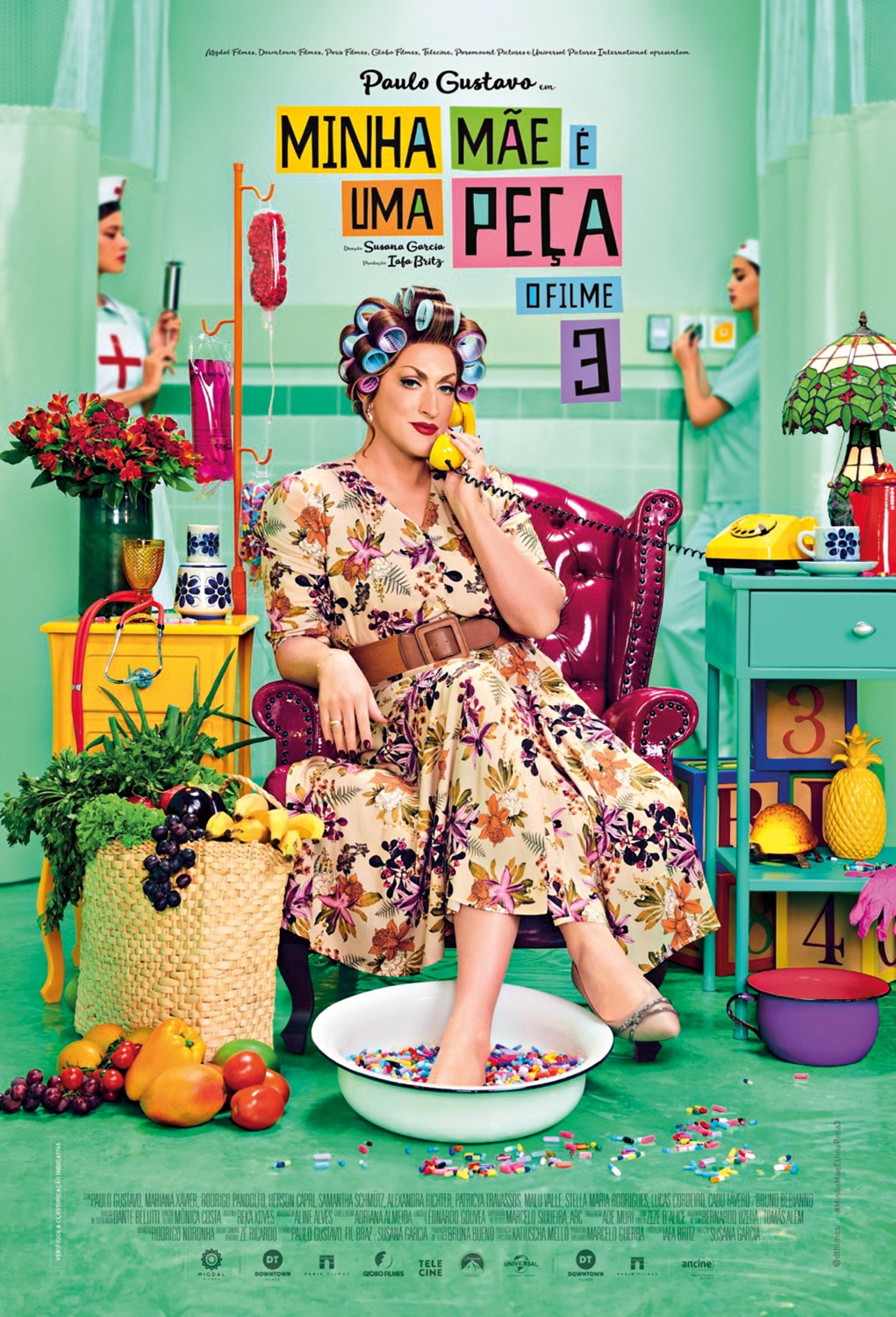 Paulo Gustavo como Dona Herm&iacute;nia, de vestido florido e bobes no cabelo, sentada em poltrona vermelha com os p&eacute;s em bacia de p&iacute;lulas coloridas, falando ao telefone amarelo. Ao fundo, enfermeiras em cen&aacute;rio hospitalar com objetos coloridos e cesta de frutas e vegetais. T&iacute;tulo: Minha M&atilde;e &Eacute; Uma Pe&ccedil;a 3 O Filme