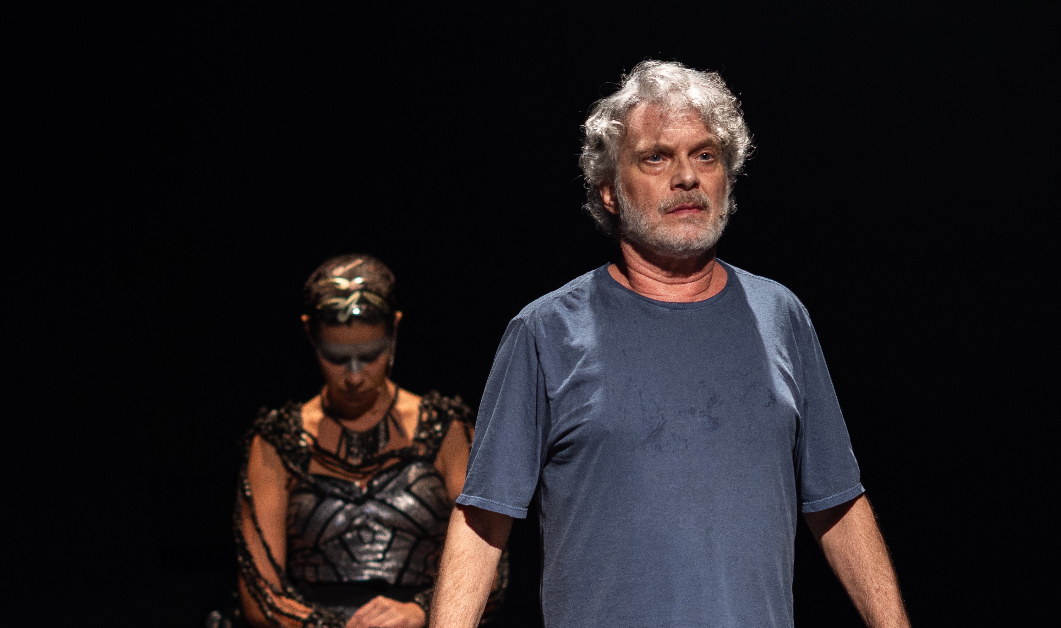 Homem de meia-idade com cabelos e barba grisalhos, vestindo camiseta azul-marinho suada, olha para frente com express&atilde;o s&eacute;ria. Atr&aacute;s dele, uma mulher com maquiagem prateada e roupa escura elaborada, de cabe&ccedil;a baixa, em um palco escuro