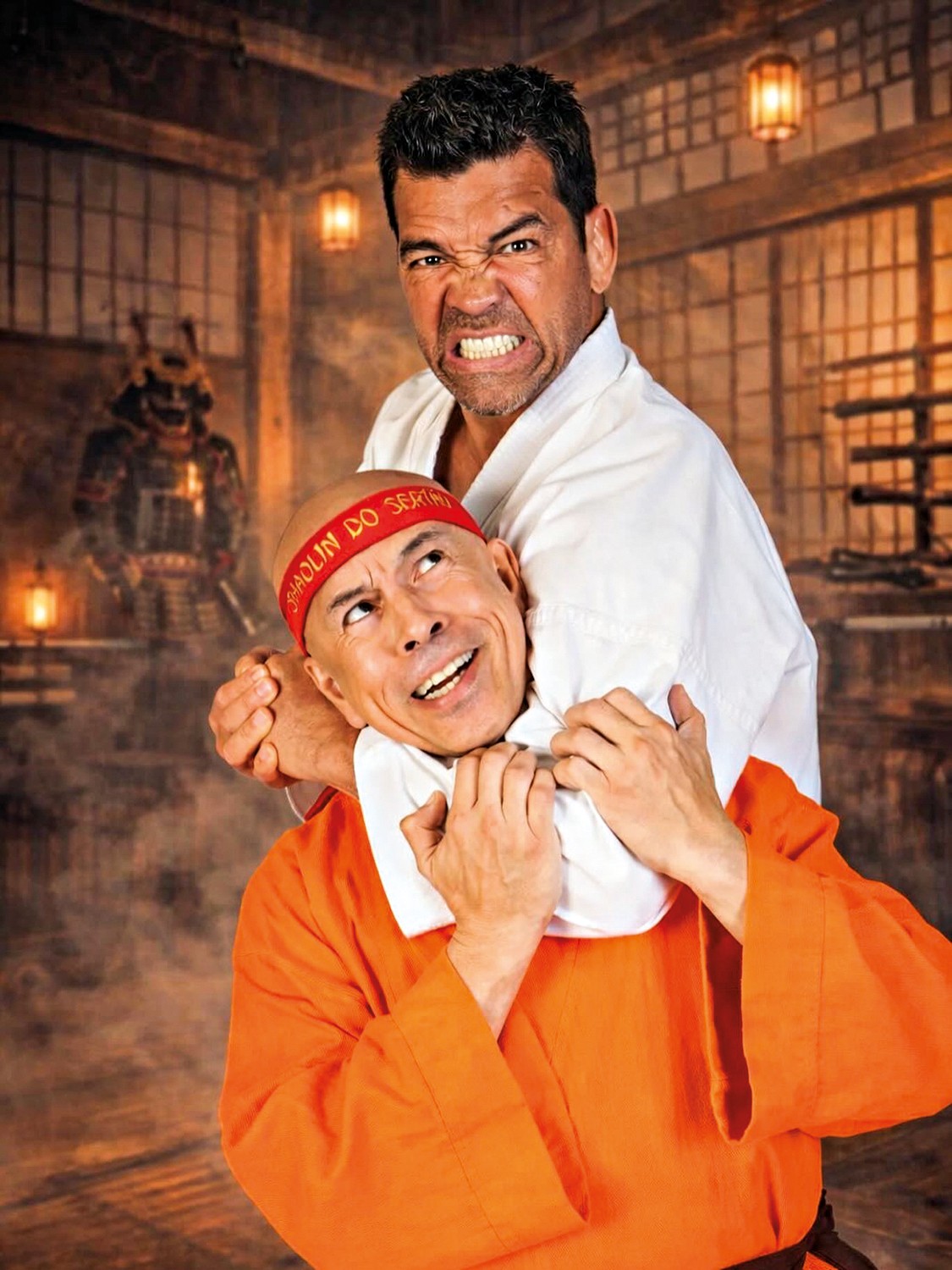 Dois homens em um dojo. Um, de quimono branco, segura o outro, de t&uacute;nica laranja e faixa vermelha Shaolin do Sert&atilde;o, pelo pesco&ccedil;o. O de branco tem express&atilde;o de raiva, dentes cerrados; o de laranja sorri, olhando para cima. Ao fundo, armadura samurai e lanternas acesas