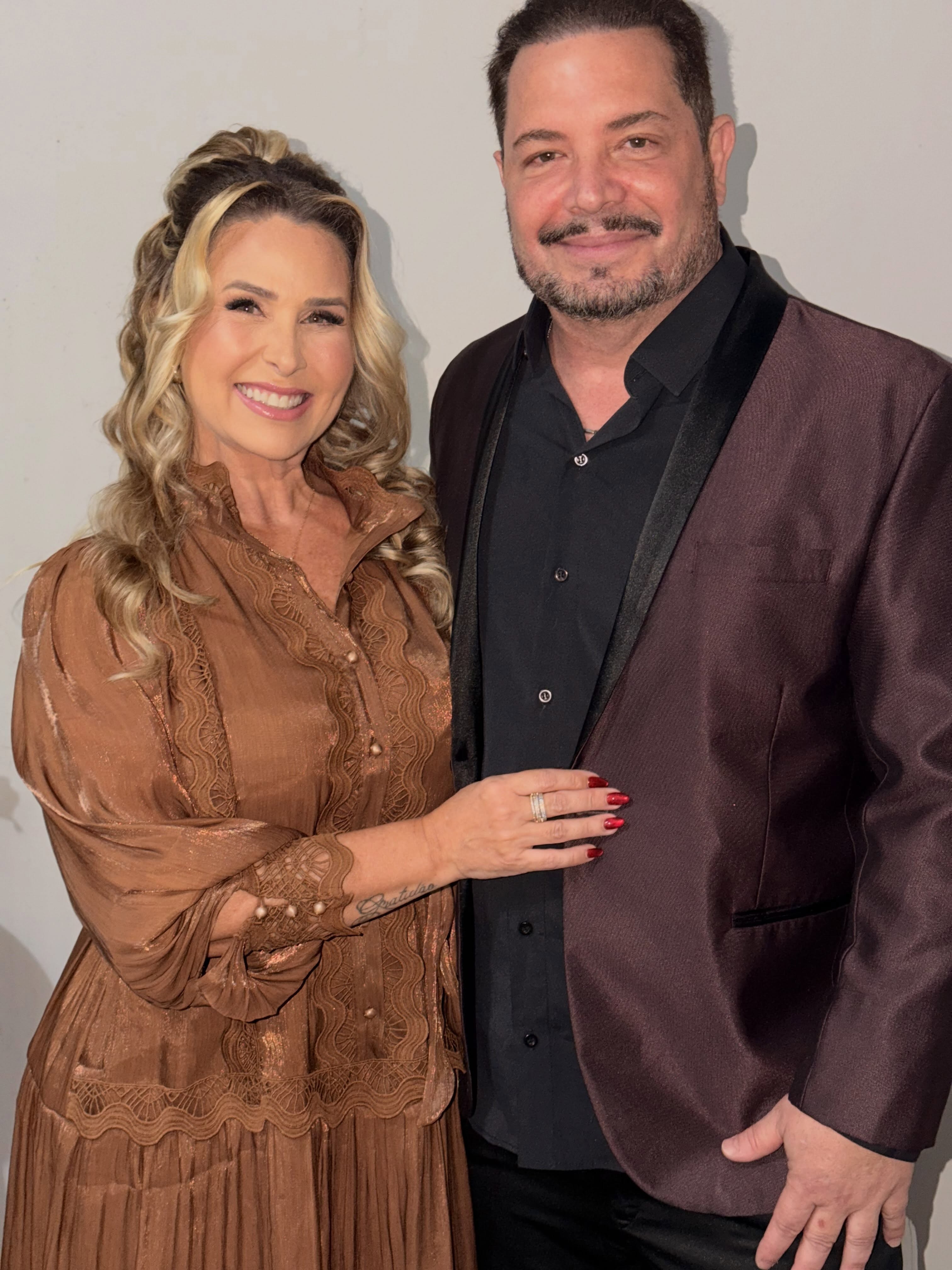 Mulher loira sorridente com vestido marrom de renda e homem barbudo com blazer vinho e camisa preta posam juntos
