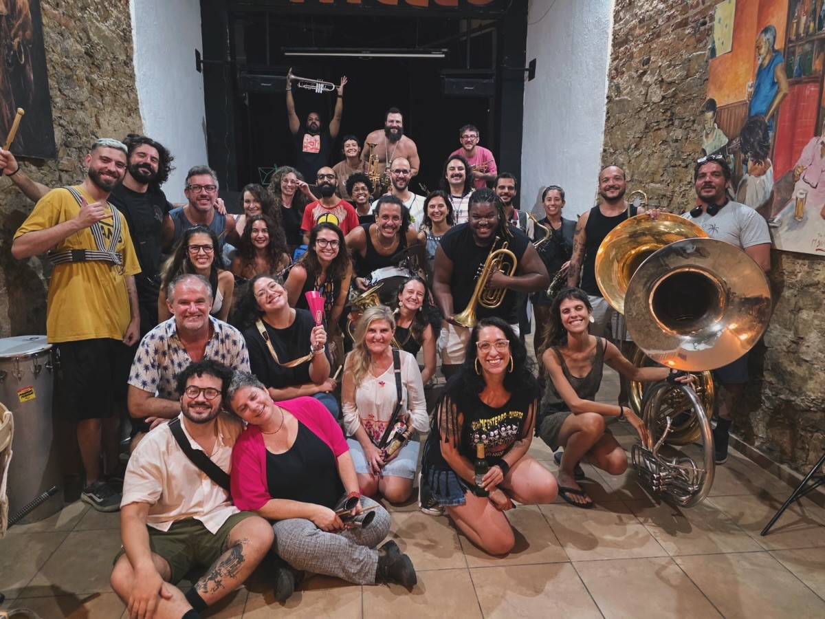 Um grande grupo de m&uacute;sicos, homens e mulheres de diversas idades, posa sorrindo para a c&acirc;mera em um ambiente interno com paredes de pedra e uma pintura colorida. Alguns seguram instrumentos de sopro, como trompetes e tubas, e outros baquetas. A maioria est&aacute; em p&eacute;, com alguns agachados na frente