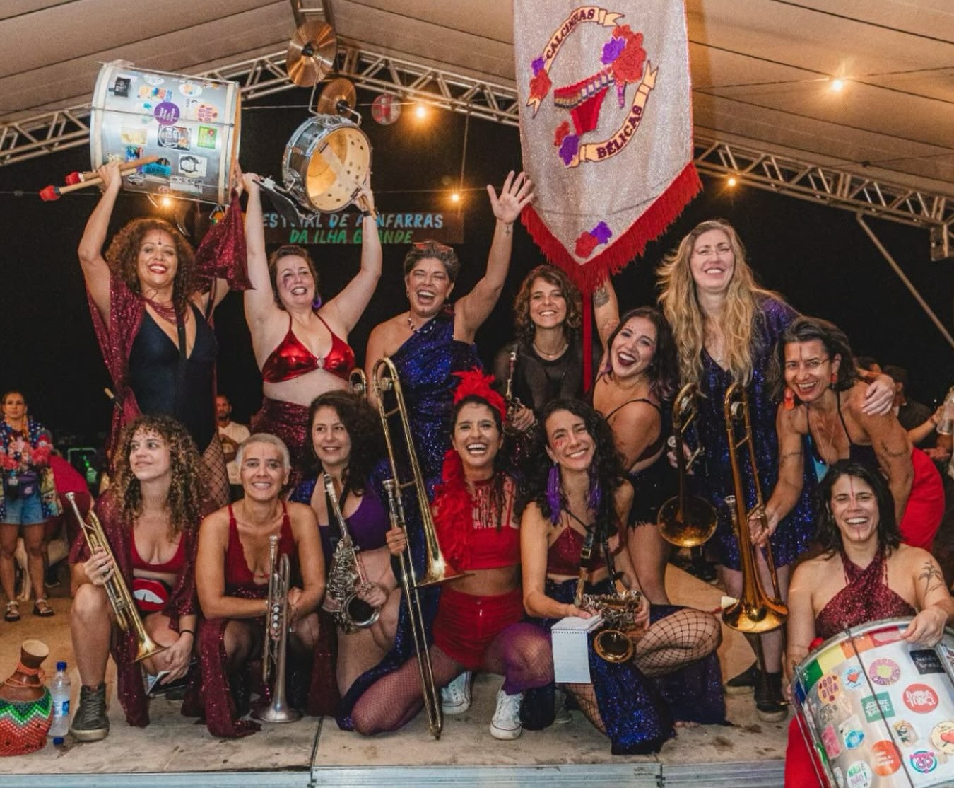 Grupo de mulheres sorrindo e posando com instrumentos musicais em um palco, algumas segurando tambores e trombones. Elas vestem roupas coloridas e brilhantes, t&iacute;picas de carnaval. Ao fundo, uma faixa com o nome CALCINHAS BELICAS e luzes penduradas. O ambiente &eacute; festivo e noturno