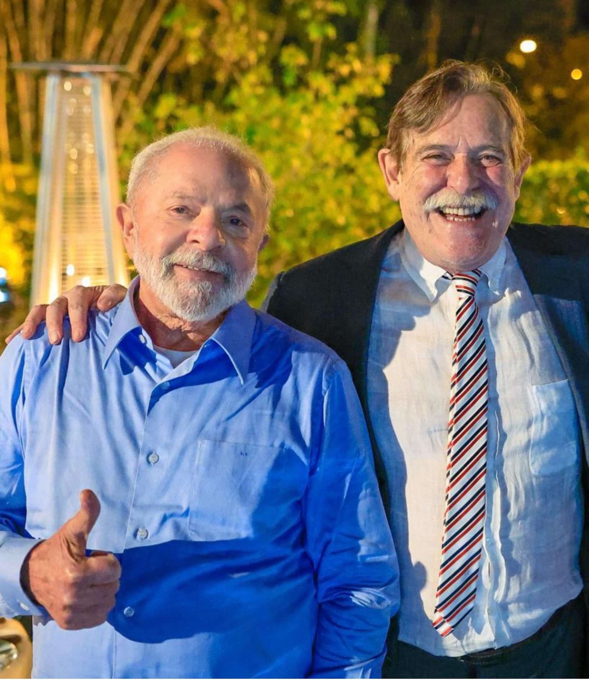 Luiz In&aacute;cio Lula da Silva, de camisa azul, sorri e faz sinal de positivo ao lado de um homem de terno e gravata listrada, que sorri amplamente. Ambos est&atilde;o em um ambiente externo com folhagens e luzes ao fundo