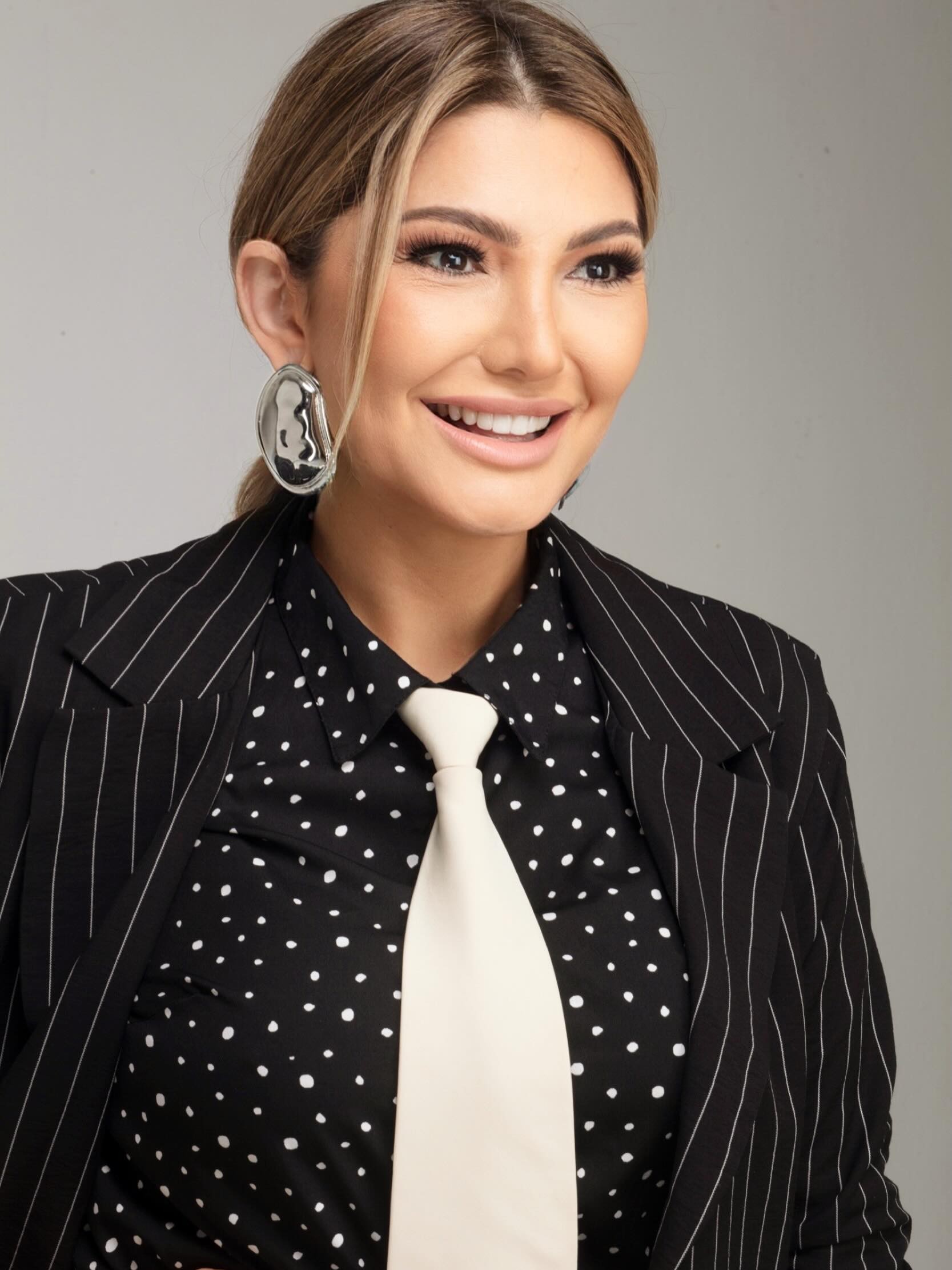 Mulher sorridente com cabelo loiro preso, maquiagem marcante, blazer preto listrado, camisa preta de bolinhas brancas e gravata creme, olhando para a direita