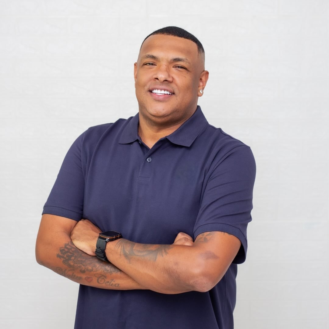 Homem negro de pele escura, sorrindo, vestindo camisa polo azul-marinho, bra&ccedil;os cruzados, rel&oacute;gio preto no pulso esquerdo e tatuagens no antebra&ccedil;o, em fundo branco