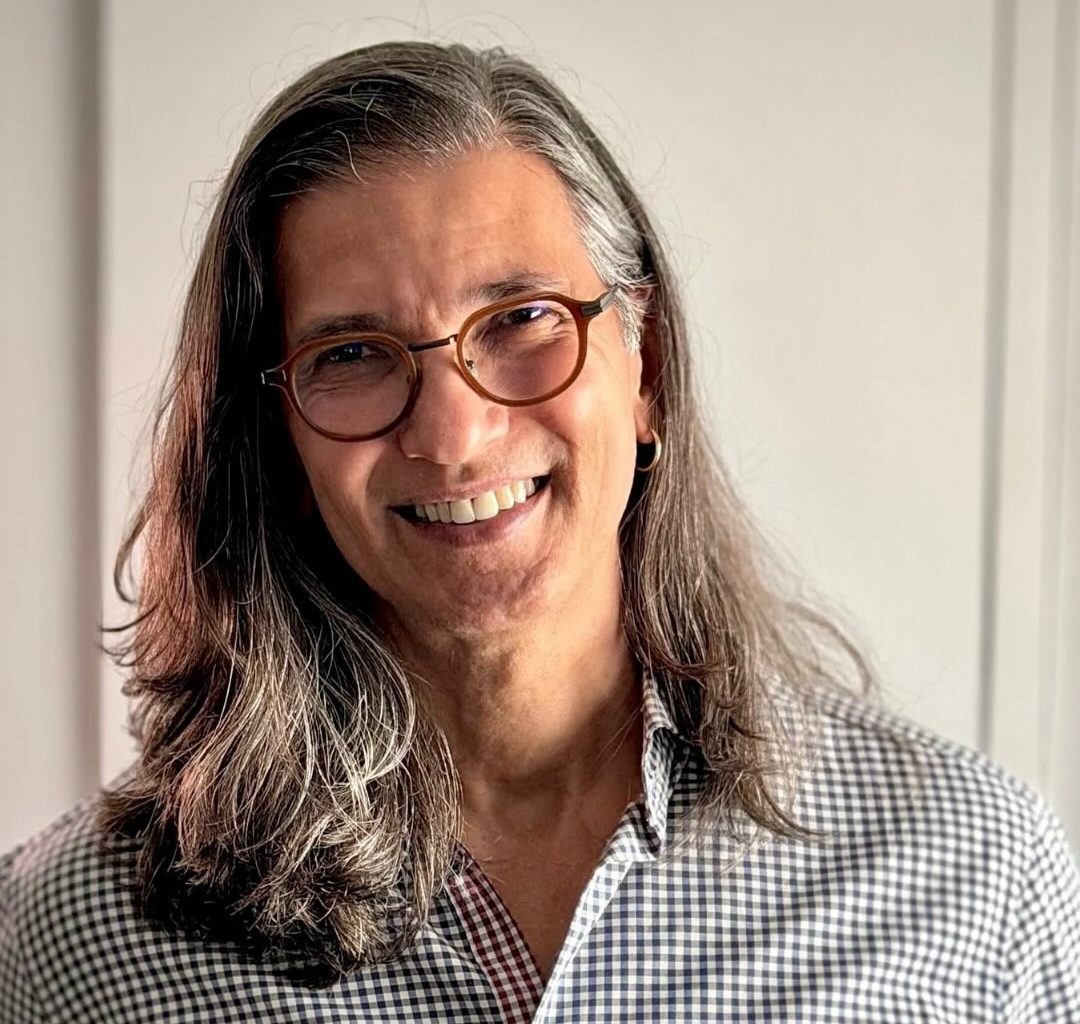 Homem de cabelos longos grisalhos, &oacute;culos redondos e camisa xadrez, sorrindo. O texto PR&Eacute;-CANDIDATURA est&aacute; em uma faixa azul no centro. Abaixo, em fundo marrom, Paulos Angelito PR&Eacute;-CANDIDATO DEPUTADO FEDERAL RJ com uma estrela estilizada &agrave; esquerda
