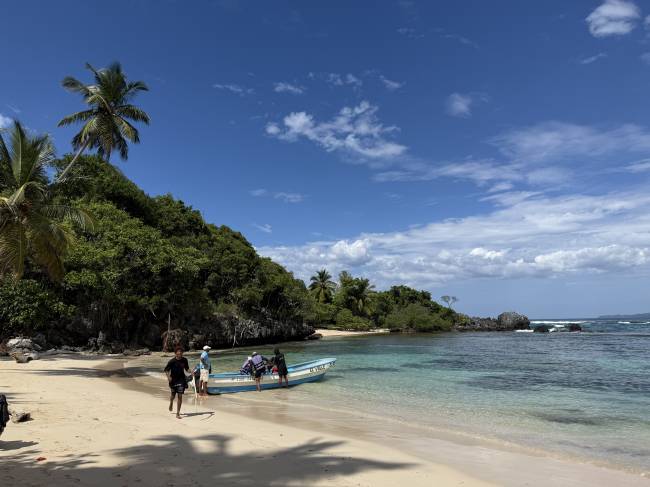 Pessoas desembarcando de um barco azul em uma praia de areia clara, com &aacute;guas cristalinas e vegeta&ccedil;&atilde;o densa na encosta, sob c&eacute;u azul com nuvens esparsas