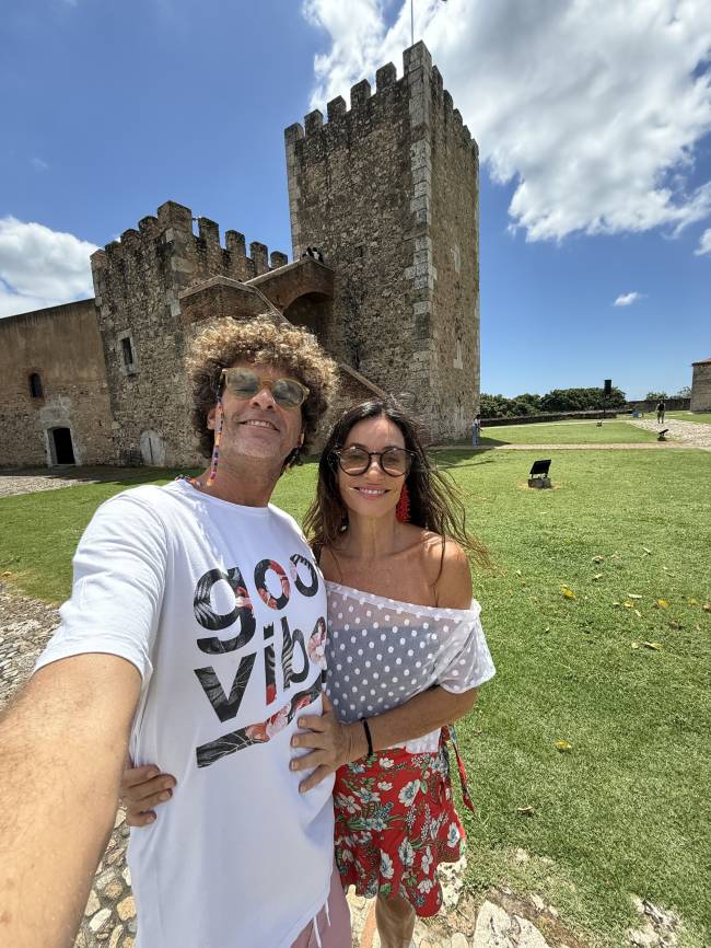 Um casal sorridente posa para selfie em frente a uma torre de pedra antiga. A mulher, de blusa branca de bolinhas e saia floral vermelha, tem cabelos longos e escuros. O homem, de camiseta branca com estampa e cabelos cacheados loiros, usa &oacute;culos de sol. Ao fundo, gramado verde e c&eacute;u azul com nuvens brancas