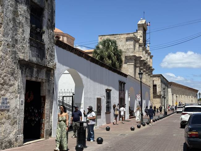 Rua hist&oacute;rica com edif&iacute;cios de pedra e paredes brancas, sob c&eacute;u azul. Pessoas caminham pela cal&ccedil;ada de tijolos, com carros estacionados &agrave; direita. Uma mulher de vestido estampado verde est&aacute; em primeiro plano, e ao fundo, uma igreja antiga com torre sineira e uma palmeira.