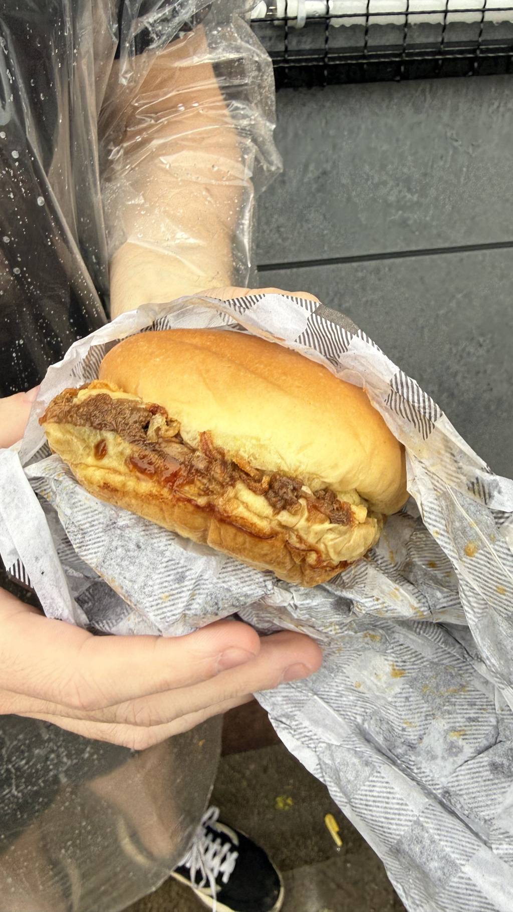 M&atilde;os segurando um sandu&iacute;che de p&atilde;o claro com recheio de carne desfiada e cebola caramelizada, envolto em papel xadrez preto e branco
