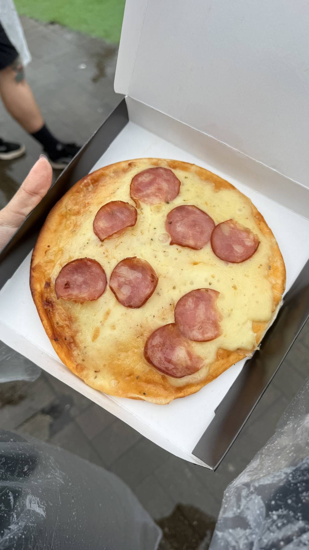Uma mini pizza de calabresa com queijo derretido e or&eacute;gano, dentro de uma caixa de papel&atilde;o branca, sendo segurada por uma m&atilde;o. Ao fundo, um ch&atilde;o molhado e a perna de uma pessoa com t&ecirc;nis pretos