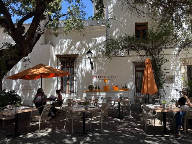 P&aacute;tio externo do Billini Hotel com mesas e cadeiras de vime, guarda-s&oacute;is laranja e um bar com garrafas. Duas mulheres conversam sob um guarda-sol e um homem bebe em outra mesa. &Aacute;rvores frondosas sombreiam o ch&atilde;o de pedras, e a fachada branca do hotel tem janelas de madeira e ferro
