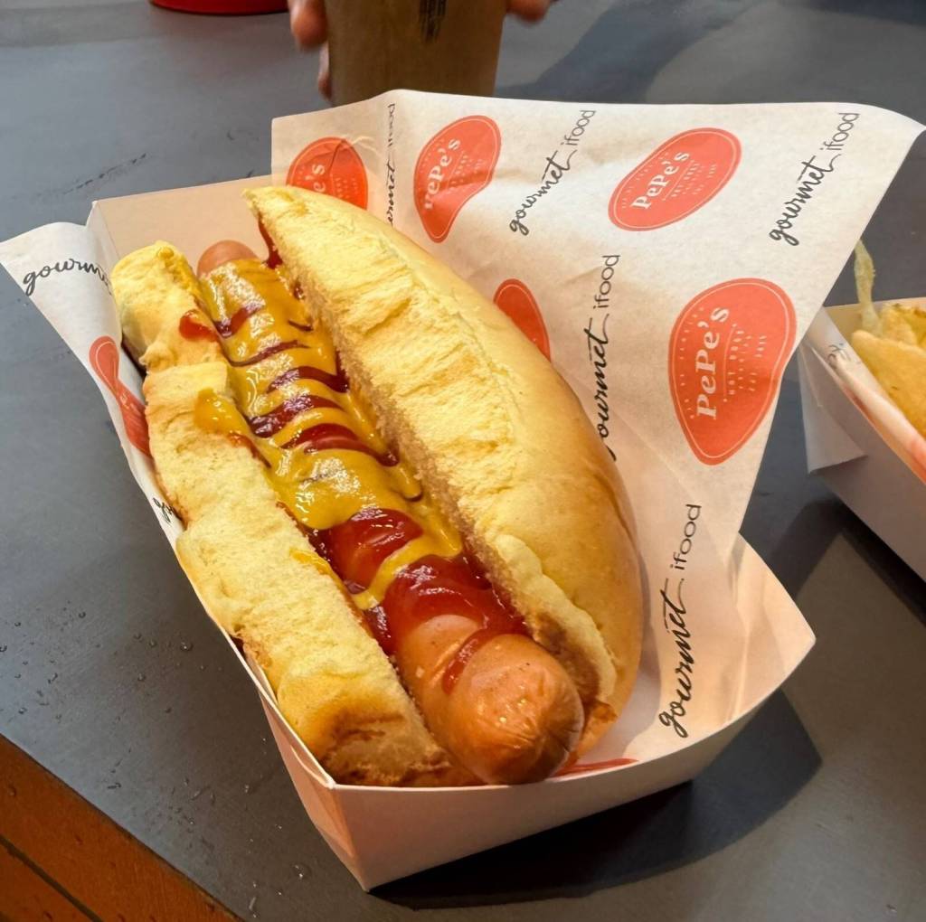 Um cachorro-quente em um p&atilde;o macio com salsicha, coberto com ketchup e mostarda, servido em uma bandeja de papel branco. O papel de embrulho ao fundo tem o logo Pepes e gourmet ifood repetidamente. Uma bebida e batatas fritas aparecem desfocadas ao fundo