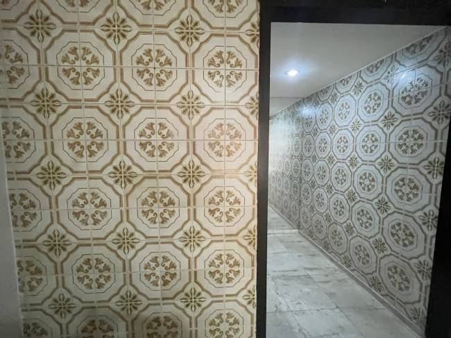 Corredor estreito com paredes revestidas de azulejos brancos e bege, com padr&otilde;es florais e geom&eacute;tricos. O piso &eacute; de m&aacute;rmore claro, e uma luz forte ilumina o teto ao fundo