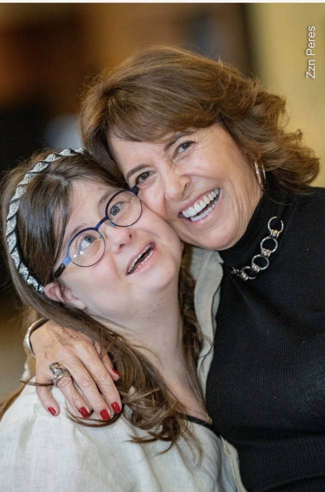 Duas mulheres sorrindo e abra&ccedil;adas. A mulher &agrave; direita, de cabelos castanhos e blusa preta, abra&ccedil;a a mulher &agrave; esquerda, de cabelos castanhos, &oacute;culos e tiara brilhante. Ambas sorriem amplamente, com a mulher da direita mostrando os dentes e a da esquerda com a boca aberta, olhando para cima. A m&atilde;o da mulher da direita, com unhas vermelhas e um anel, repousa no ombro da outra