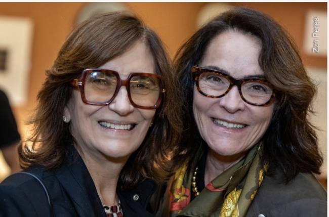 Duas mulheres de meia-idade, sorrindo para a c&acirc;mera. A da esquerda usa &oacute;culos grandes marrons e brincos de p&eacute;rola. A da direita usa &oacute;culos de tartaruga e um len&ccedil;o estampado verde e dourado