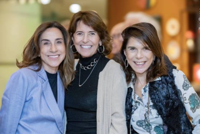 Tr&ecirc;s mulheres sorridentes, de meia-idade, posam lado a lado. A da esquerda veste blazer azul-claro sobre blusa preta. A do centro usa blusa preta de gola alta, colar prateado e casaco bege. A da direita veste blusa estampada com colete de pele sint&eacute;tica preto.