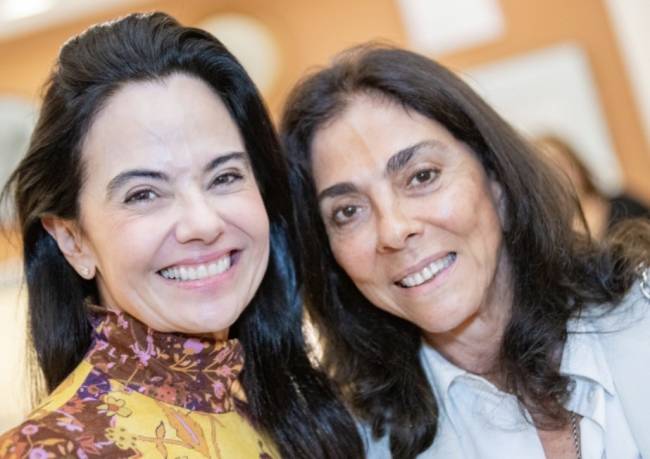 Duas mulheres sorridentes, de cabelos escuros, posam lado a lado. A da esquerda veste blusa amarela com estampa floral marrom e roxa, enquanto a da direita usa camisa clara