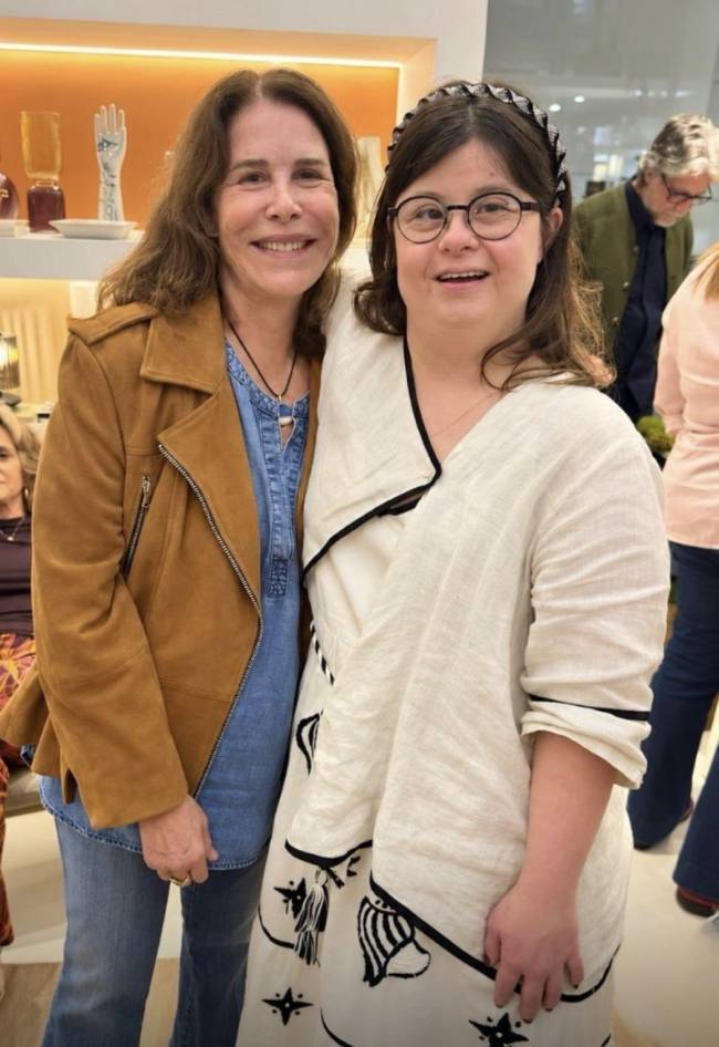 Duas mulheres sorrindo para a c&acirc;mera. Uma mulher de cabelos castanhos e jaqueta marrom abra&ccedil;a uma jovem com &oacute;culos e tiara preta, vestindo um casaco branco com detalhes pretos. Ao fundo, pessoas e prateleiras com objetos decorativos.