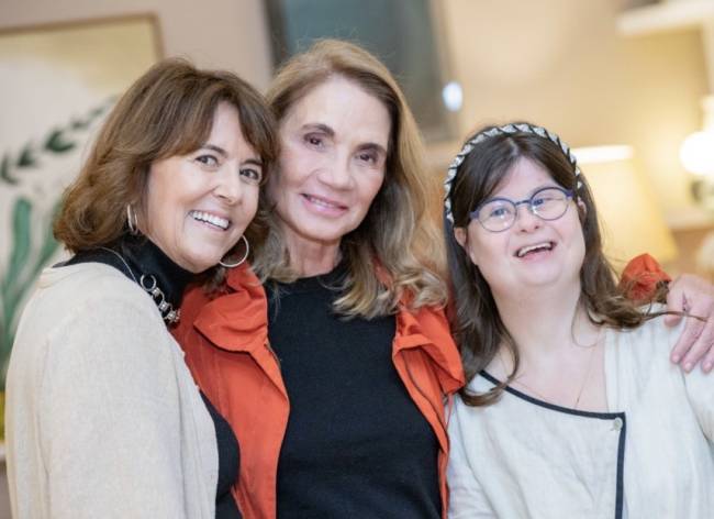 Tr&ecirc;s mulheres sorridentes posam juntas, a da esquerda com cabelo castanho e blusa bege, a do centro com cabelo loiro e blusa preta com jaqueta laranja, e a da direita, com s&iacute;ndrome de Down, &oacute;culos redondos e tiara, vestindo blusa branca com detalhes pretos