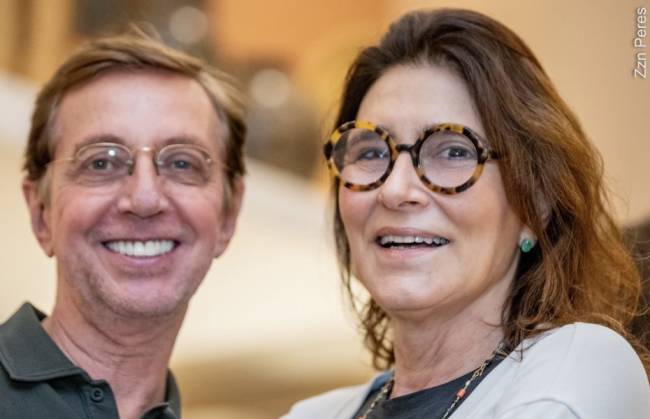 Um homem sorridente com &oacute;culos e camisa polo verde-escura ao lado de uma mulher sorridente com &oacute;culos de tartaruga e brinco verde