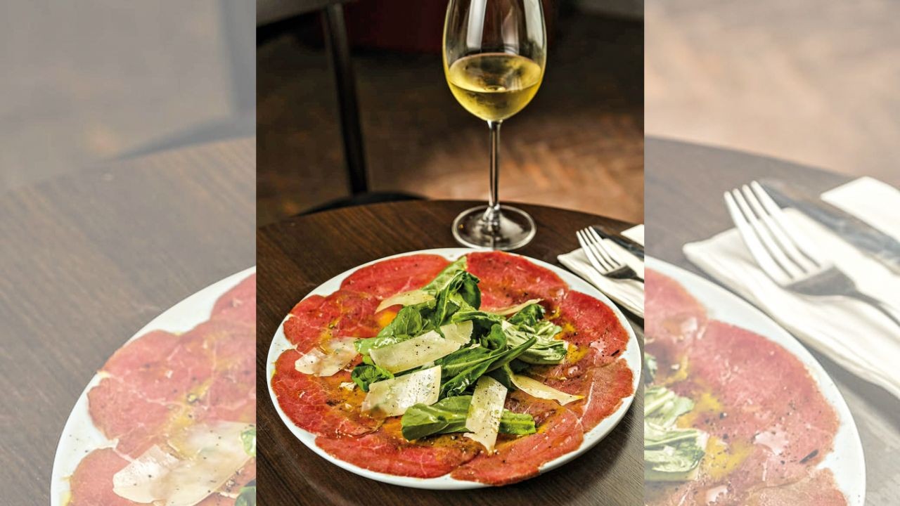 Carpaccio de carne com folhas verdes, lascas de queijo parmesão e molho, servido em prato branco sobre mesa de madeira escura, com taça de vinho branco e talheres ao lado