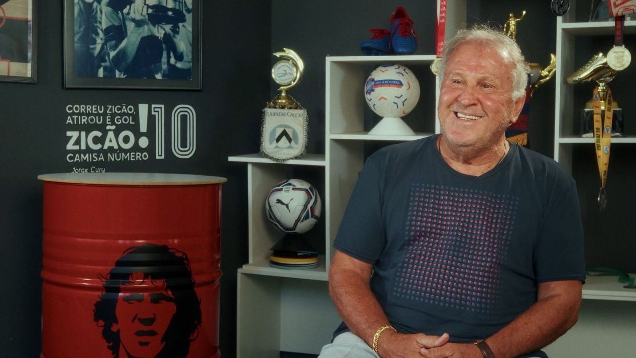 Um homem idoso de pele clara, cabelos grisalhos e sorriso largo, vestindo camiseta azul escura e calça branca, sentado em um ambiente com prateleiras cheias de troféus e bolas de futebol. À esquerda, um barril vermelho com a silhueta de um rosto e uma frase sobre Zicão