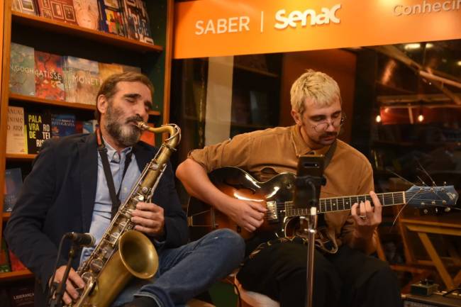 Dois m&uacute;sicos tocam instrumentos: um homem barbudo de camisa azul e blazer escuro toca saxofone, e um jovem de cabelo loiro e &oacute;culos redondos toca viol&atilde;o el&eacute;trico. Eles est&atilde;o sentados em um ambiente com estantes de livros ao fundo e um letreiro laranja com SABER | Senac acima