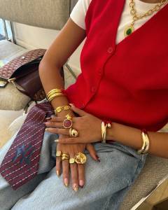 Pessoa sentada com blusa branca, colete vermelho e cal&ccedil;a jeans, exibindo v&aacute;rias joias douradas: colares, pulseiras e an&eacute;is grandes. As unhas t&ecirc;m esmalte dourado e detalhes em vermelho. Uma bolsa marrom com monograma est&aacute; ao lado