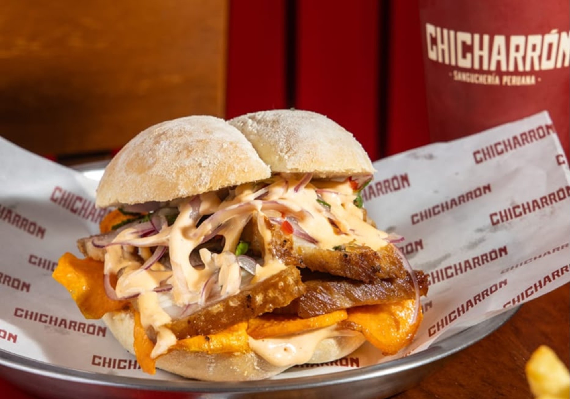 Sanduíche de chicharrón peruano com pão crocante, carne de porco frita, batata doce laranja, cebola roxa fatiada e molho cremoso, servido em papel com o nome Chicharrón em um prato metálico, com um copo vermelho ao fundo
