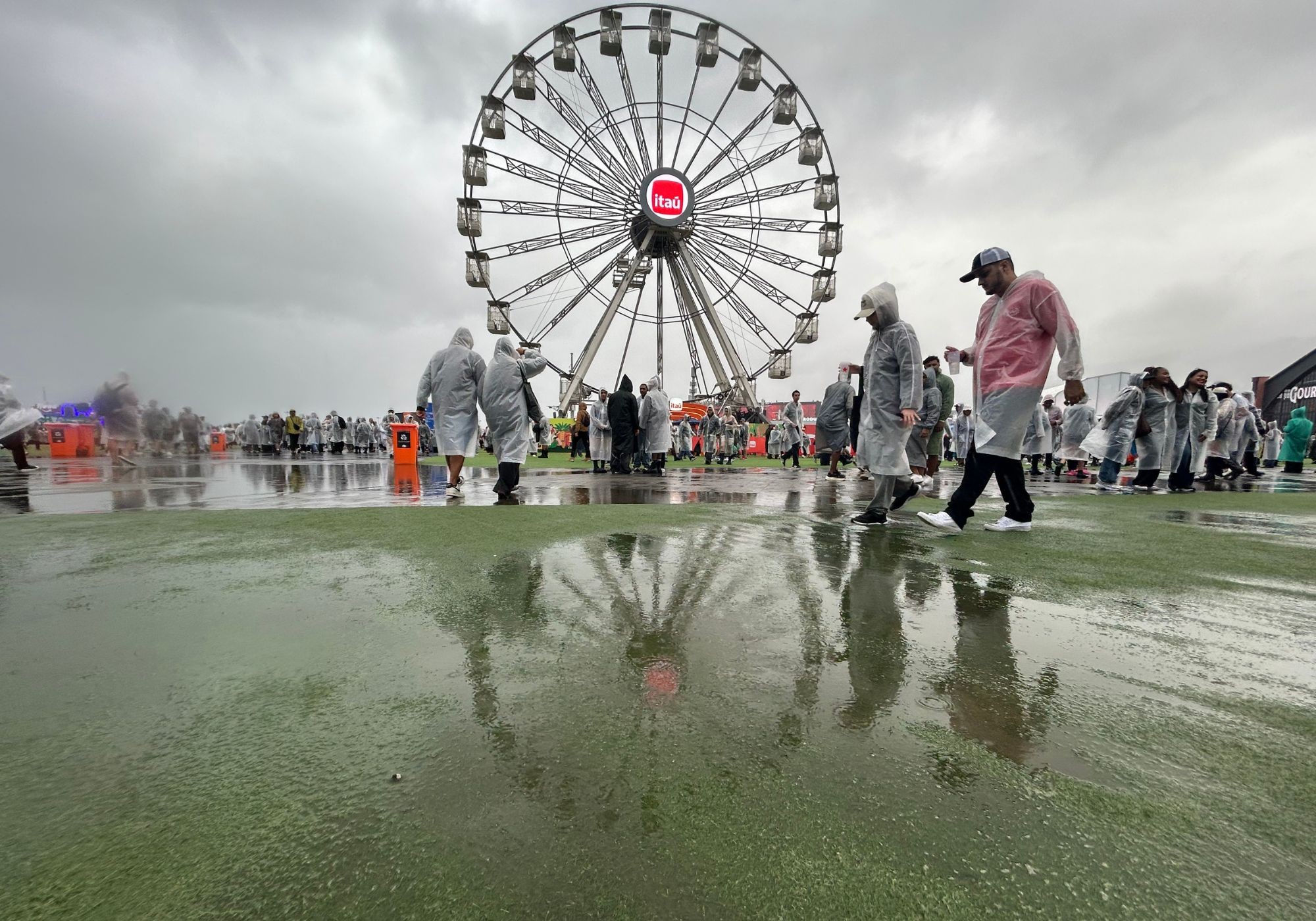 Pessoas com capas de chuva transparentes caminham sobre gramado molhado, refletindo uma roda-gigante com o logo Itaú sob céu nublado