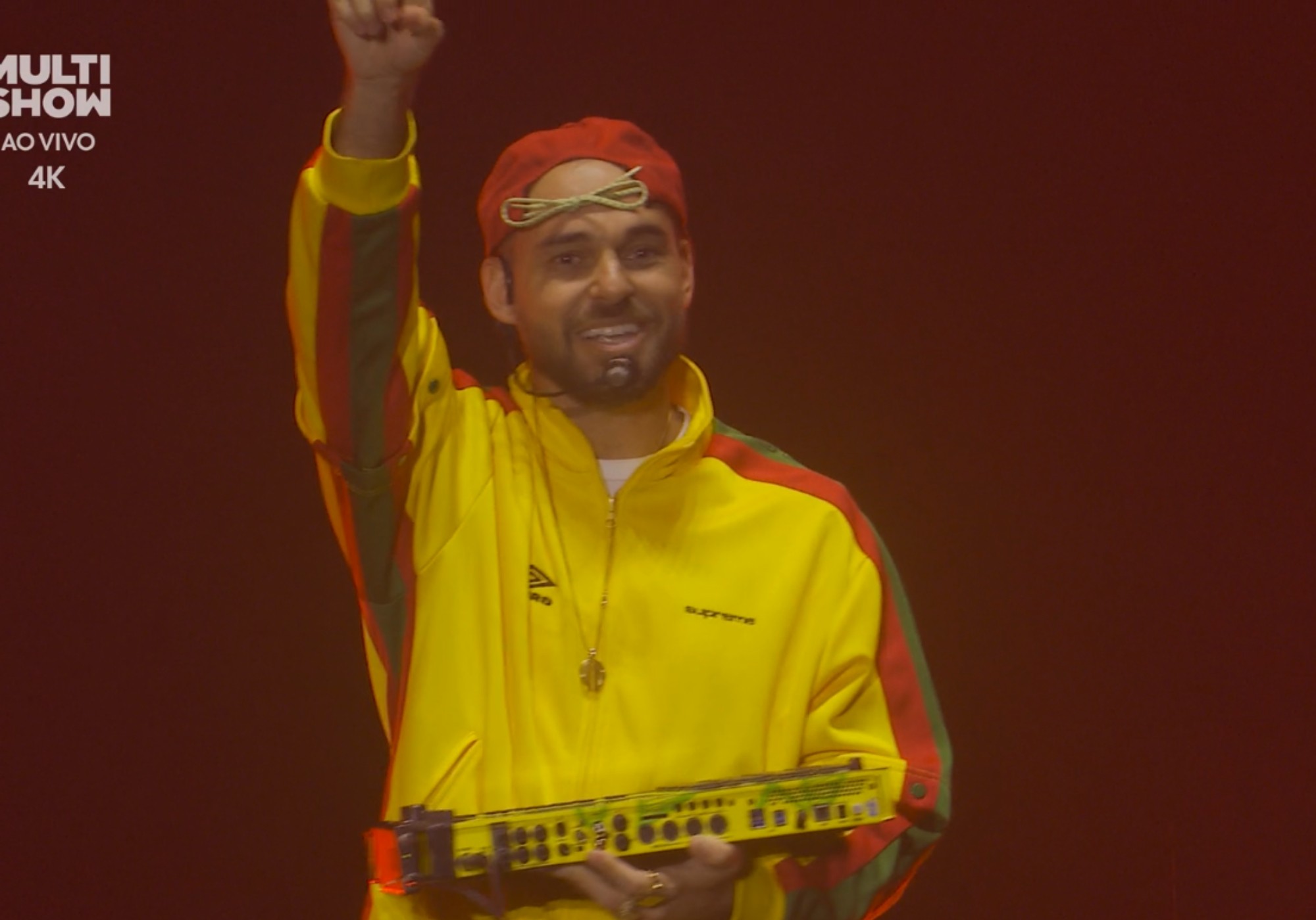 Homem sorridente com barba, boné vermelho e jaqueta amarela com listras vermelhas e verdes, segurando um teclado musical amarelo e levantando o punho direito, em palco escuro