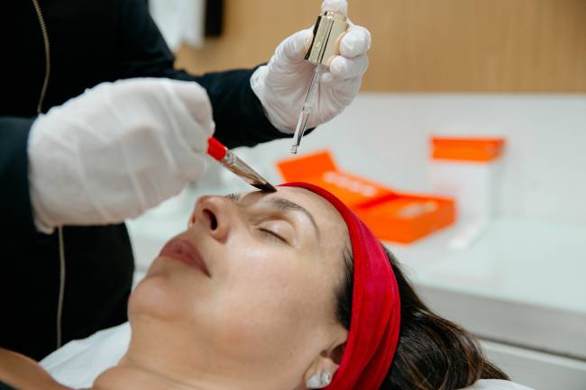 Uma mulher deitada com os olhos fechados e uma faixa de cabelo vermelha, recebendo um tratamento facial. Uma m&atilde;o com luva branca aplica um produto na testa dela com um pincel, enquanto a outra m&atilde;o segura um conta-gotas com s&eacute;rum.