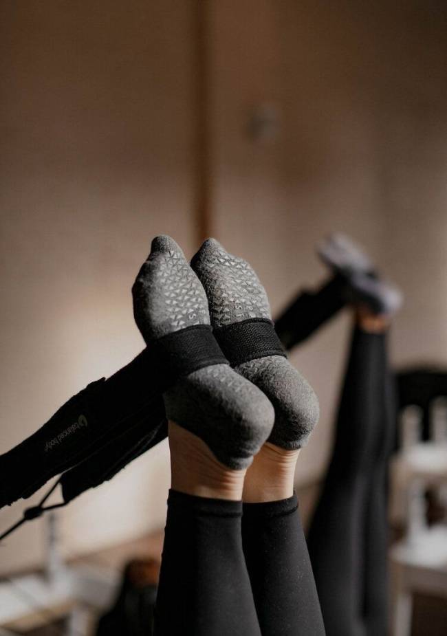 P&eacute;s de uma pessoa usando meias cinzas antiderrapantes e cal&ccedil;a legging preta, presos a uma al&ccedil;a preta de equipamento de pilates com o logo Balanced Body, em um est&uacute;dio