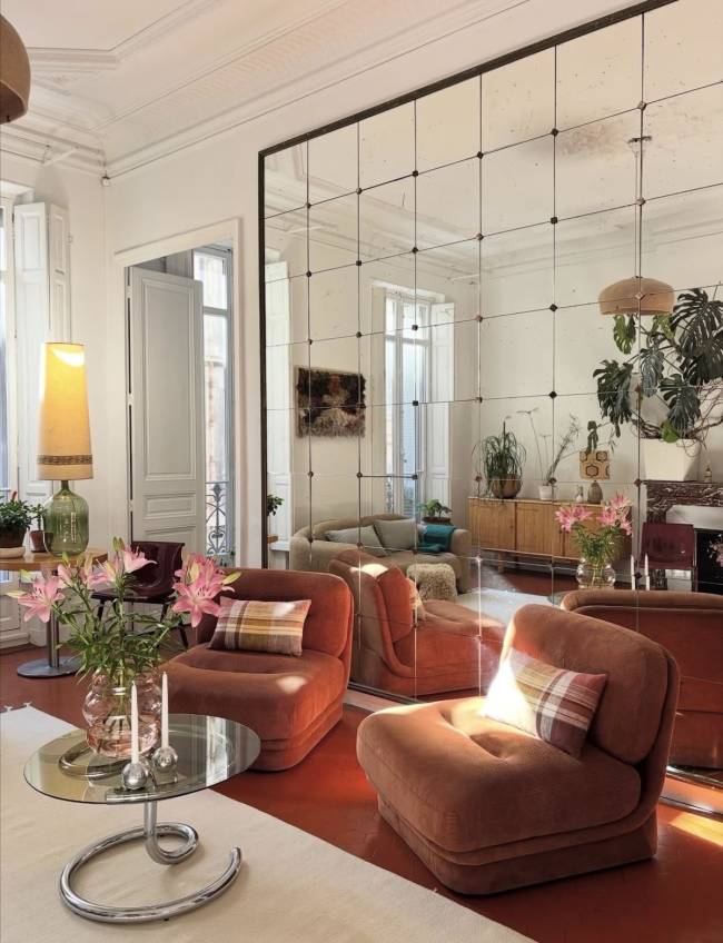 Sala de estar com poltronas estofadas cor de telha, almofadas xadrez, mesa de centro de vidro com flores cor-de-rosa e velas. Um grande espelho quadriculado reflete o ambiente, incluindo um sof&aacute; e plantas. O ch&atilde;o &eacute; de cor telha e h&aacute; um tapete claro. Uma lumin&aacute;ria alta com c&uacute;pula amarela est&aacute; &agrave; esquerda