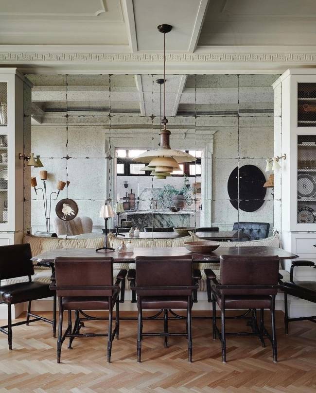 Sala de jantar com piso de madeira em espinha de peixe, mesa retangular escura com cadeiras de couro marrom e um banco estofado. A parede ao fundo &eacute; coberta por espelhos envelhecidos, refletindo um ambiente com plantas e uma lumin&aacute;ria pendente de design moderno