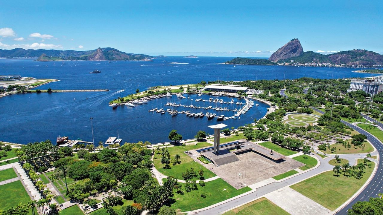 Vista aérea da Baía de Guanabara, Rio de Janeiro, com o Pão de Açúcar ao fundo. Em primeiro plano, o Monumento aos Pracinhas, um parque verdejante e uma marina com diversos barcos atracados. O céu é azul claro, e a água, azul escura.