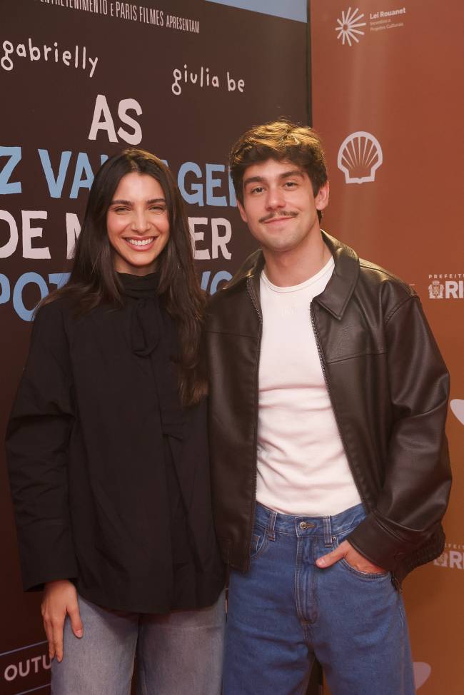 Uma mulher e um homem jovens sorriem para a c&acirc;mera, em p&eacute; diante de um painel de divulga&ccedil;&atilde;o. Ela tem cabelos longos e escuros, veste blusa preta de gola alta. Ele tem bigode, cabelo castanho e veste jaqueta de couro marrom sobre camiseta branca, com cal&ccedil;a jeans. O painel ao fundo tem textos e logotipos de patrocinadores