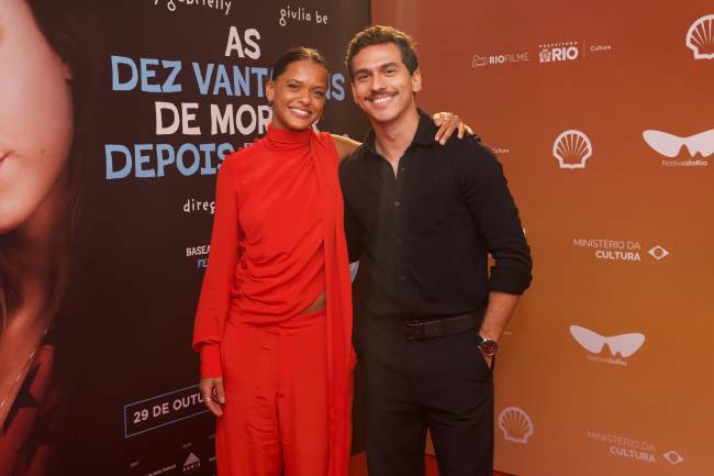 Uma mulher negra de blusa vermelha de gola alta e cal&ccedil;a larga vermelha sorri ao lado de um homem branco de camisa preta e cal&ccedil;a preta, com o bra&ccedil;o sobre o ombro dela. Ambos est&atilde;o em um evento, com um painel de fundo escuro e outro laranja com logotipos