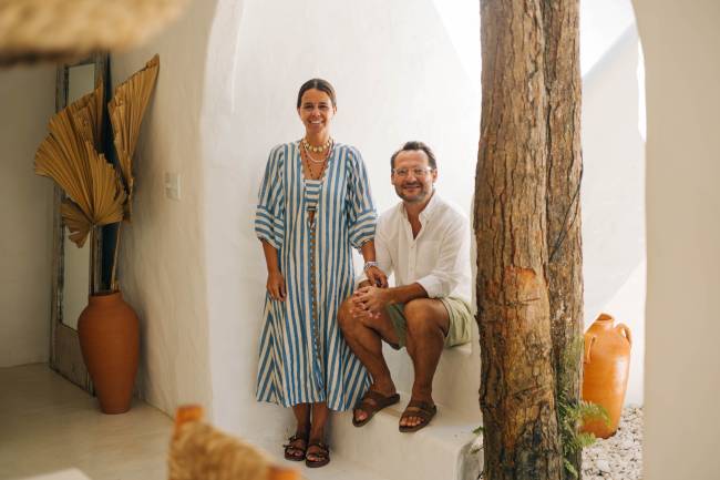 Mulher sorridente de vestido listrado azul e branco ao lado de homem de camisa branca e bermuda, sentado em degrau branco, em ambiente claro com tronco de &aacute;rvore e vasos de barro